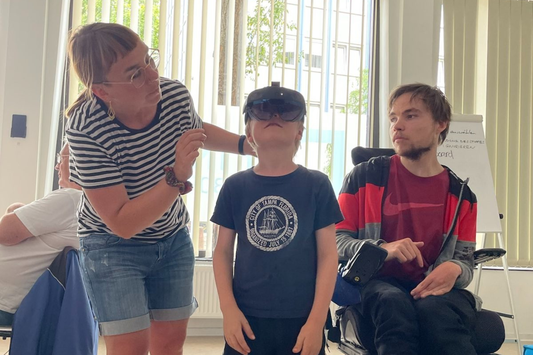 Frau setzt einem Jungen eine Virtual-Reality-Brille auf, daneben sitzt ein junger Mann im Rollstuhl
