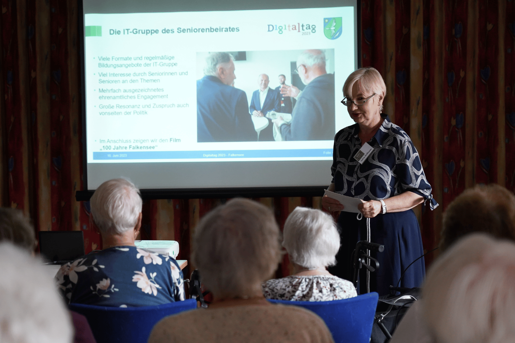 Ein ältere Frau spricht vor einer Gruppe Menschen, im Hintergrung eine Leinwand, auf der eine Präsentation zum Seniorenbeirat Falkensee projiziert wird