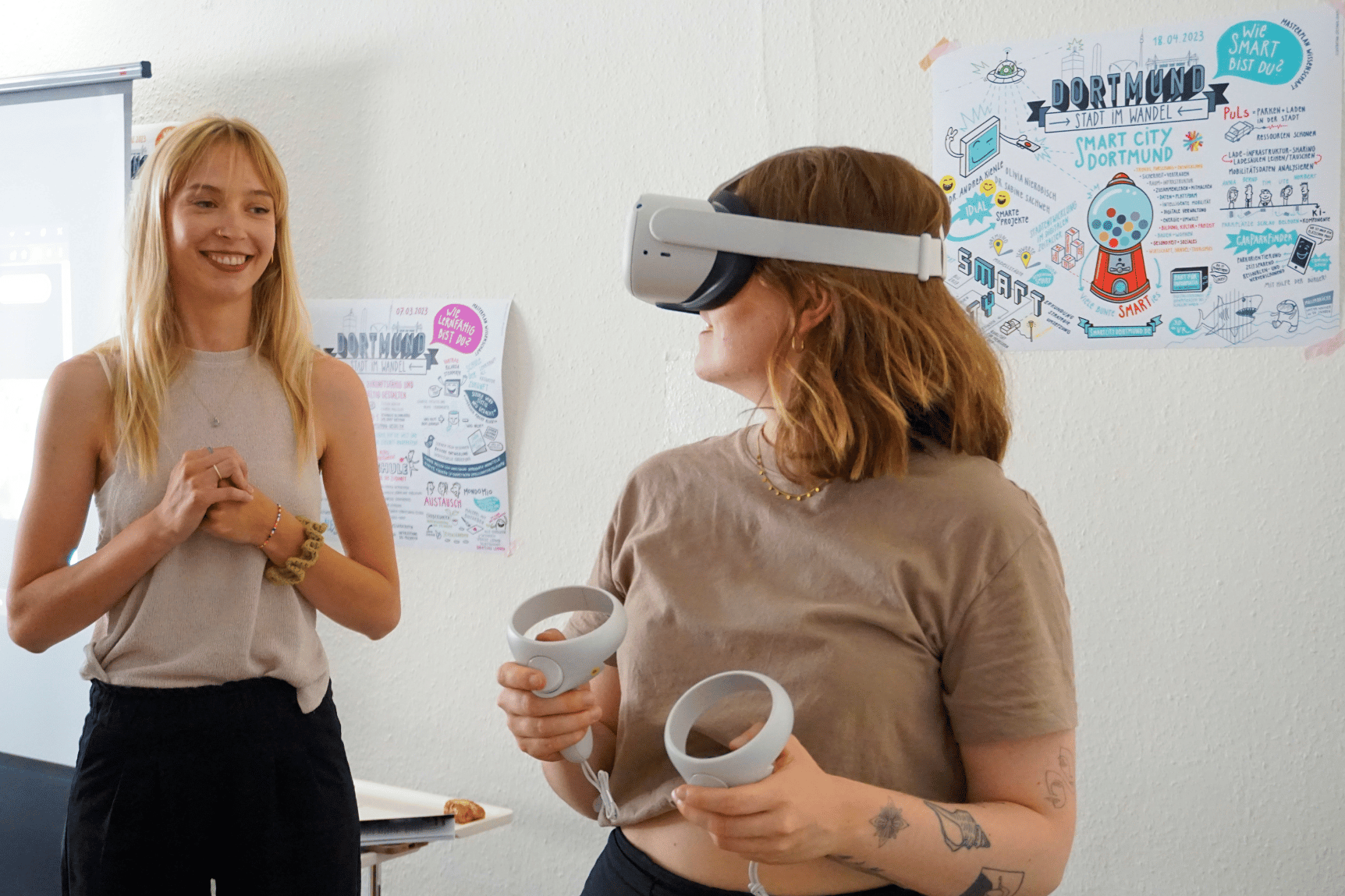 Zwei junge Frauen, die, die rechts steht, trägt eine VR-Brille und hält zwei Controller in den Händen