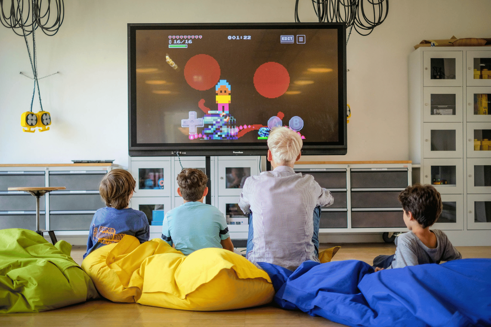 Ein Mann und drei Kinder sitzen auf Sitzsäcken auf dem Boden und schauen sich ein Videospiel auf einem großen Fernseher an, der an der Wand hängt