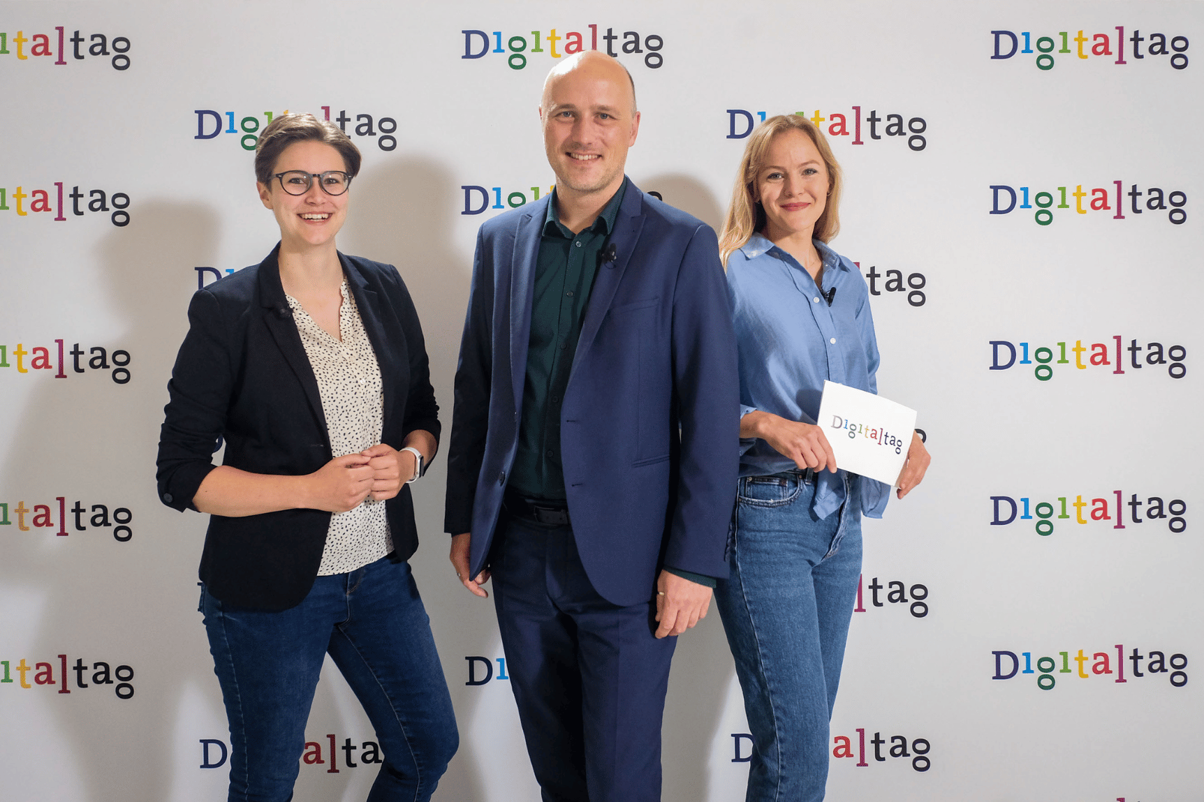 Staatssekretär Sven Lehmann steht mit den zwei Moderatorinnen des Digitaltag-Livestreams vor einer Digitaltag-Fotowand