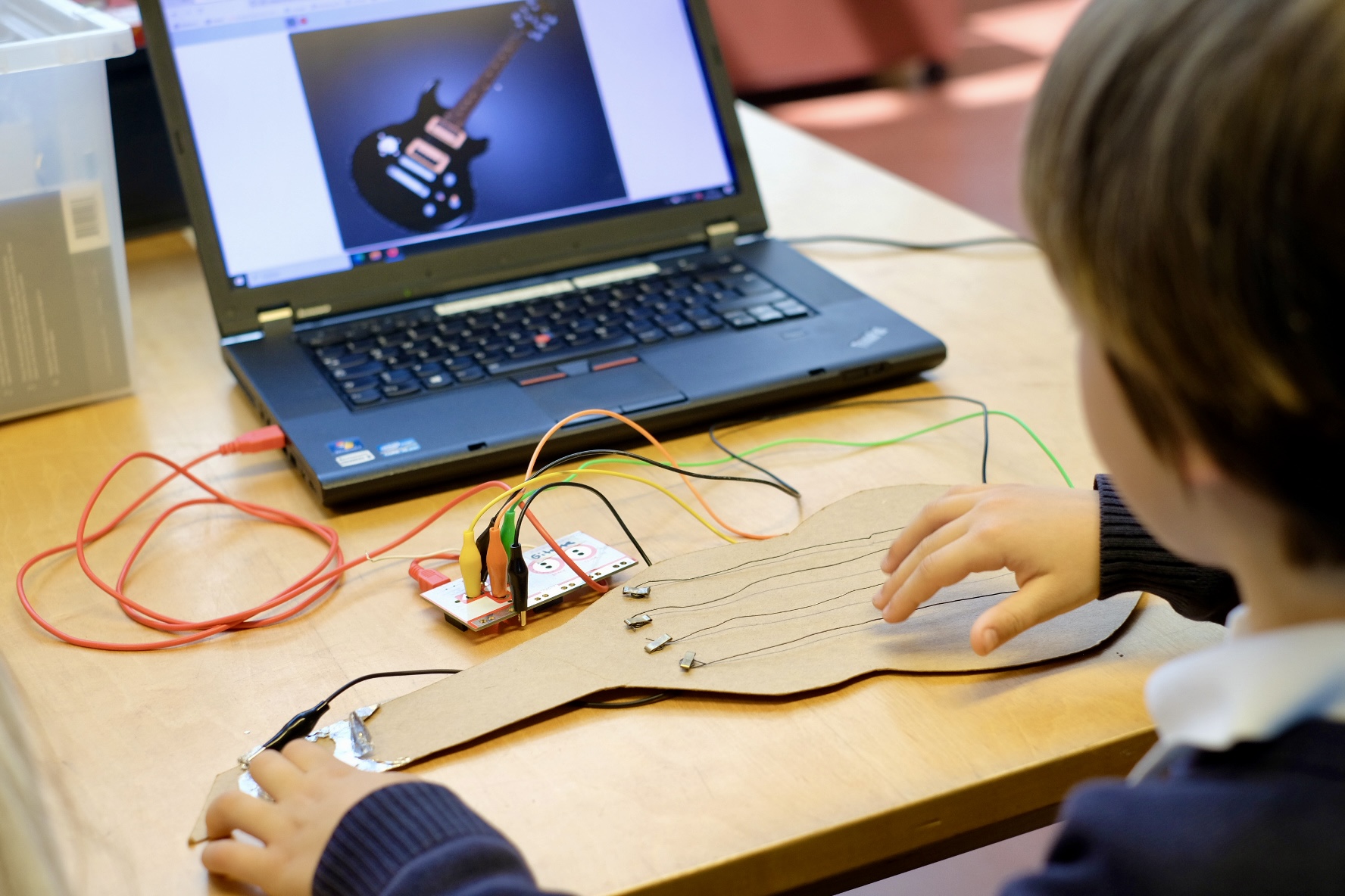 Das Bild zeigt die Hände eines Kindes, das an einem elektronischen Projekt arbeitet. Es beinhaltet einen Kartonausschnitt mit leitfähigen Pfaden und verschiedenen Drähten, die an eine kleine Platine angeschlossen sind. Diese Platine ist wiederum mit einem Laptop verbunden, auf dem ein Bild einer Gitarre angezeigt wird. Es scheint sich um eine Bildungsaktivität zu handeln, die möglicherweise mit dem Erlernen von Elektronik, Leitfähigkeit oder Computerprogrammierung in einem Klassenzimmer zusammenhängt. 