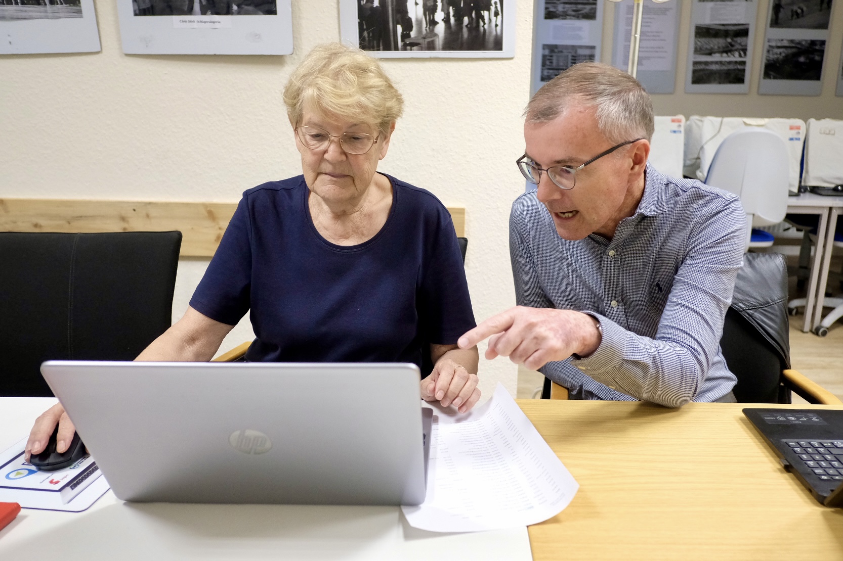 Das Bild zeigt zwei Personen, die an einem Tisch sitzen und über einige Papiere diskutieren, die auf dem Tisch liegen. Vor der Person im blauen Oberteil steht ein geöffneter Laptop. Neben dem Laptopbenutzer sind ein Taschenrechner und weitere Papiere zu sehen.