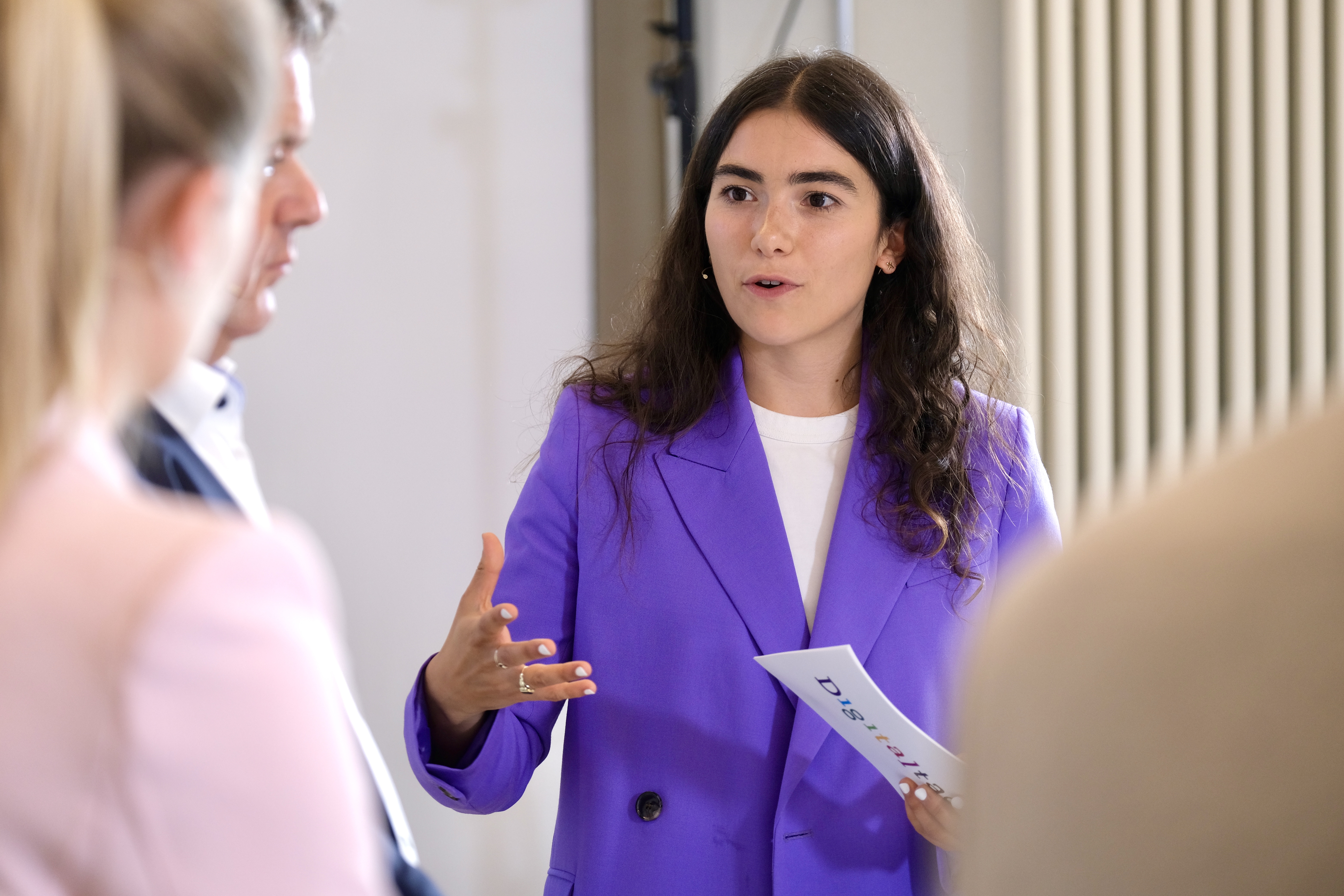 Das Bild zeigt drei Personen. Die zentrale Figur scheint mit einer Hand zu gestikulieren, während sie in der anderen eine Moderationskarte hält. Diese Person trägt einen lila Blazer. Die anderen beiden Figuren sind der zentralen Figur zugewandt und nur von hinten zu sehen.