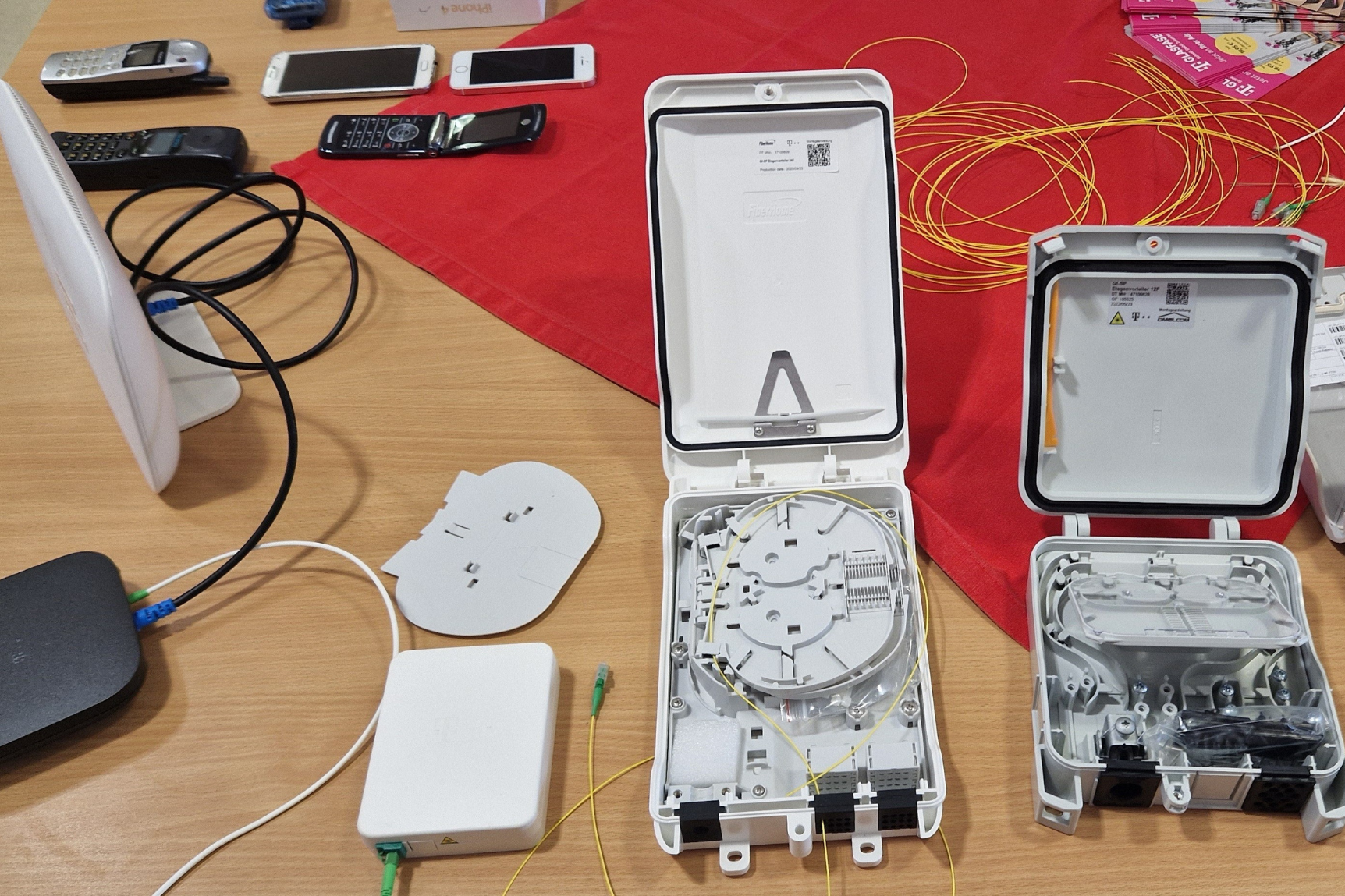 Das Bild zeigt eine Vielzahl von elektronischen Geräten und Komponenten, die auf einem Tisch ausgebreitet sind. Zu sehen sind zwei offene Gehäuse mit elektronischen Teilen darin, Kabel und mehrere Mobiltelefone. 