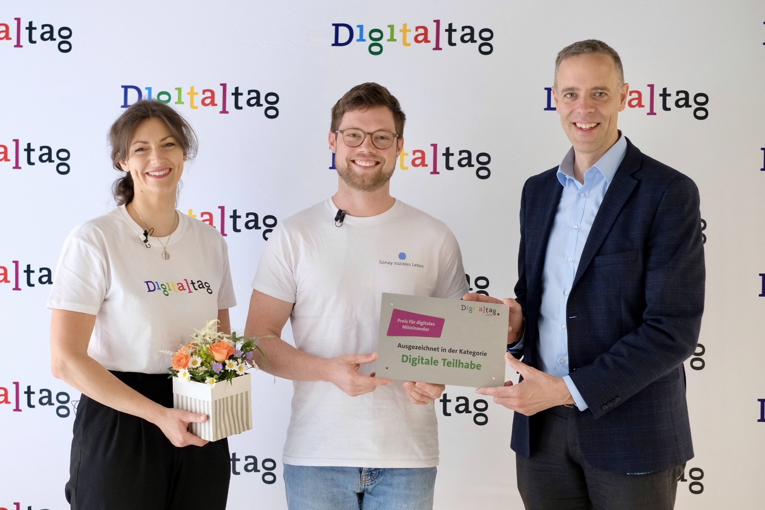 Foto des Preisträgers des Preises für digitales Miteinander 2024 mit der Moderatorin des Livestreams, die ein Digitaltag-T-Shirt trägt, und Dr. Markus Richter von der Digitaltag-Fotowand.