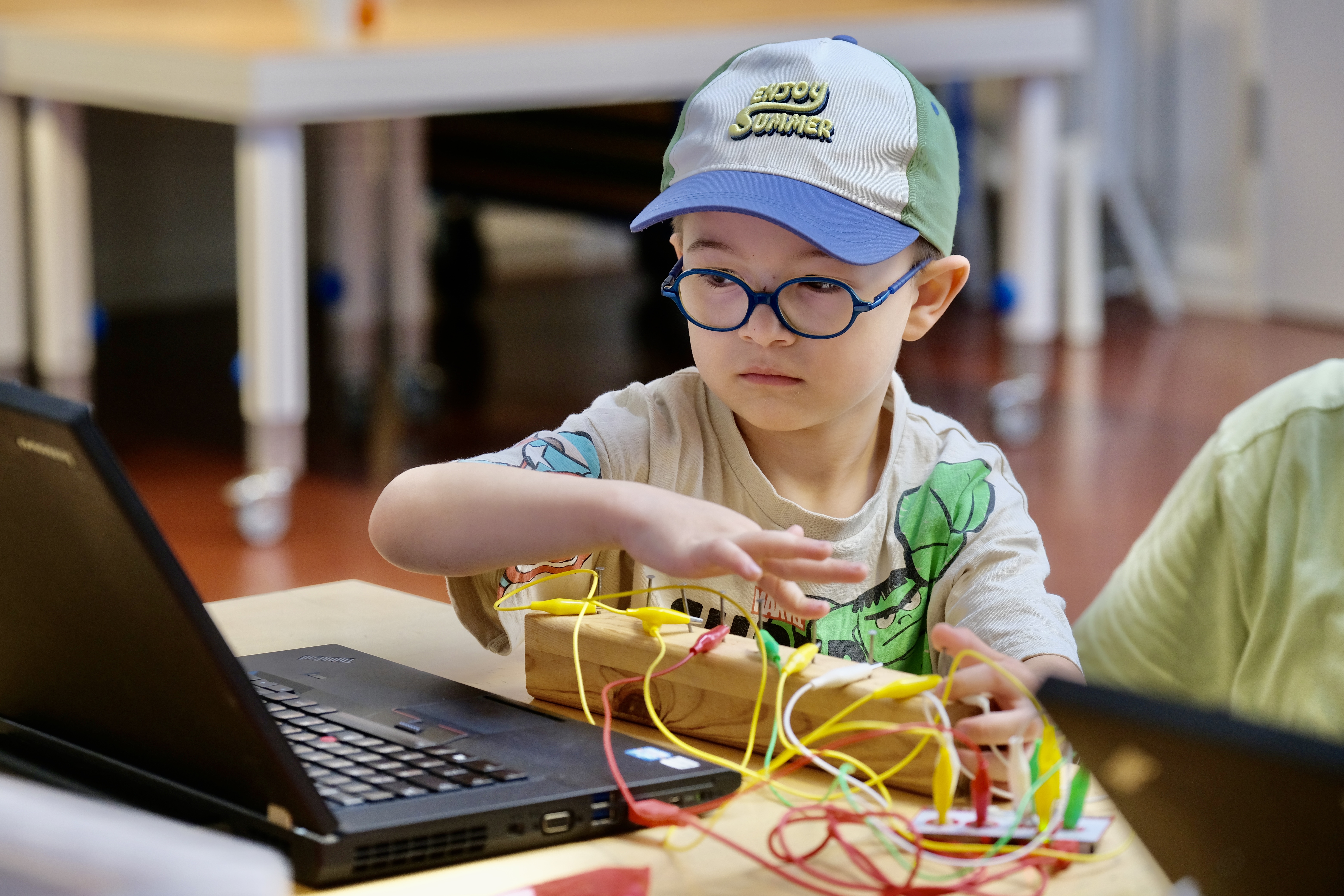 Ein kleiner Junge mit Brille sitzt vor einem Laptop und einem länglichen Holzblock, zu dem mehrere bunte Kabel führen. 