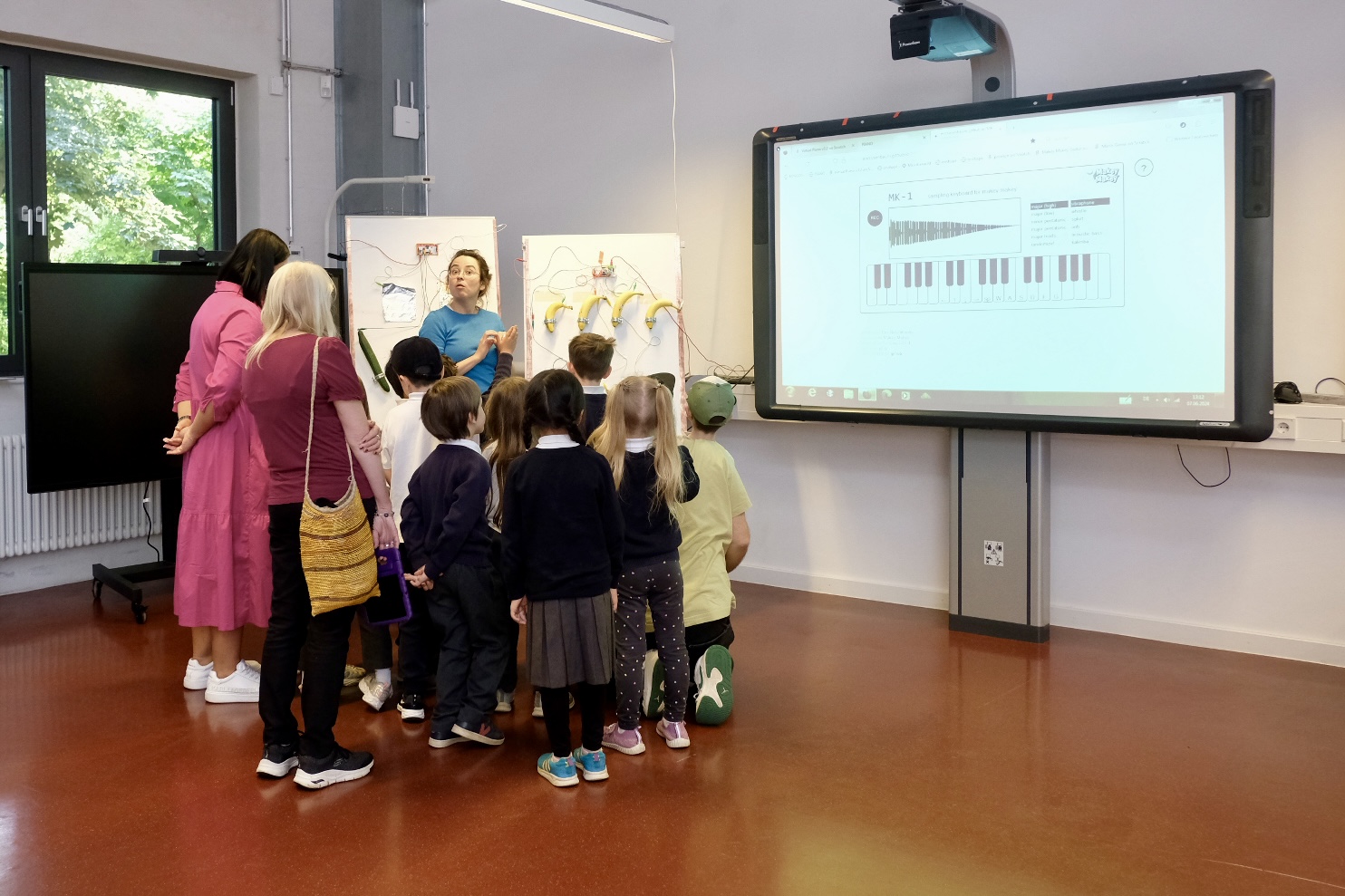 Das Bild zeigt eine Gruppe von Kindern und zwei Erwachsenen in einem Klassenzimmer, die sich auf einen großen Bildschirm an der Vorderseite des Raumes konzentrieren. Auf dem Bildschirm ist eine Klaviertastatur zu sehen. 