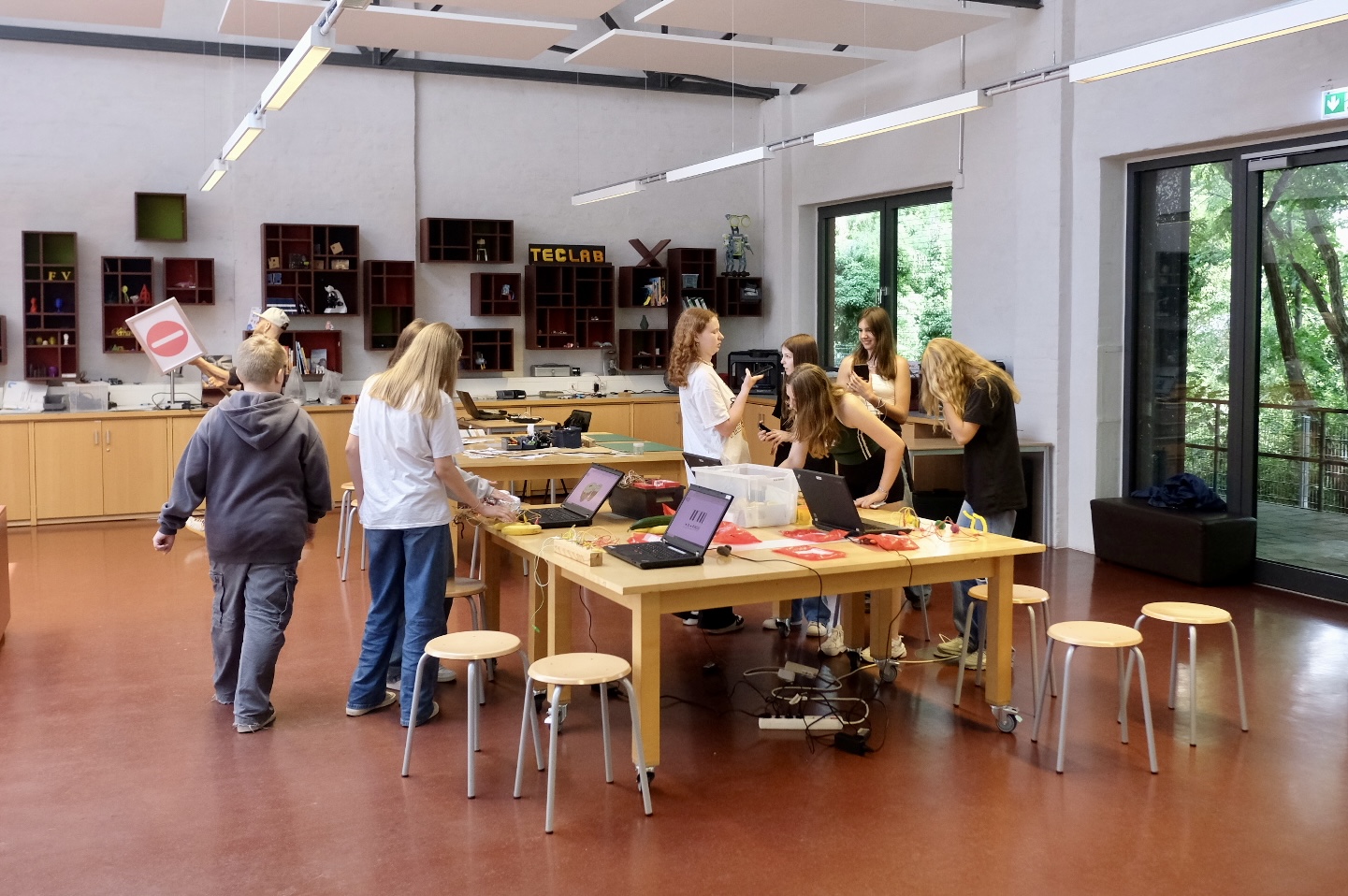 Das Bild zeigt eine Klassenzimmerumgebung mit mehreren Personen, die um einen großen Tisch stehen. Es gibt Hocker um den Tisch herum, und der Raum ist mit Labor- oder technischem Equipment ausgestattet. Die Wände haben Fenster, die auf Grünflächen hinausblicken, und an der Decke befinden sich Leuchtstofflampen.