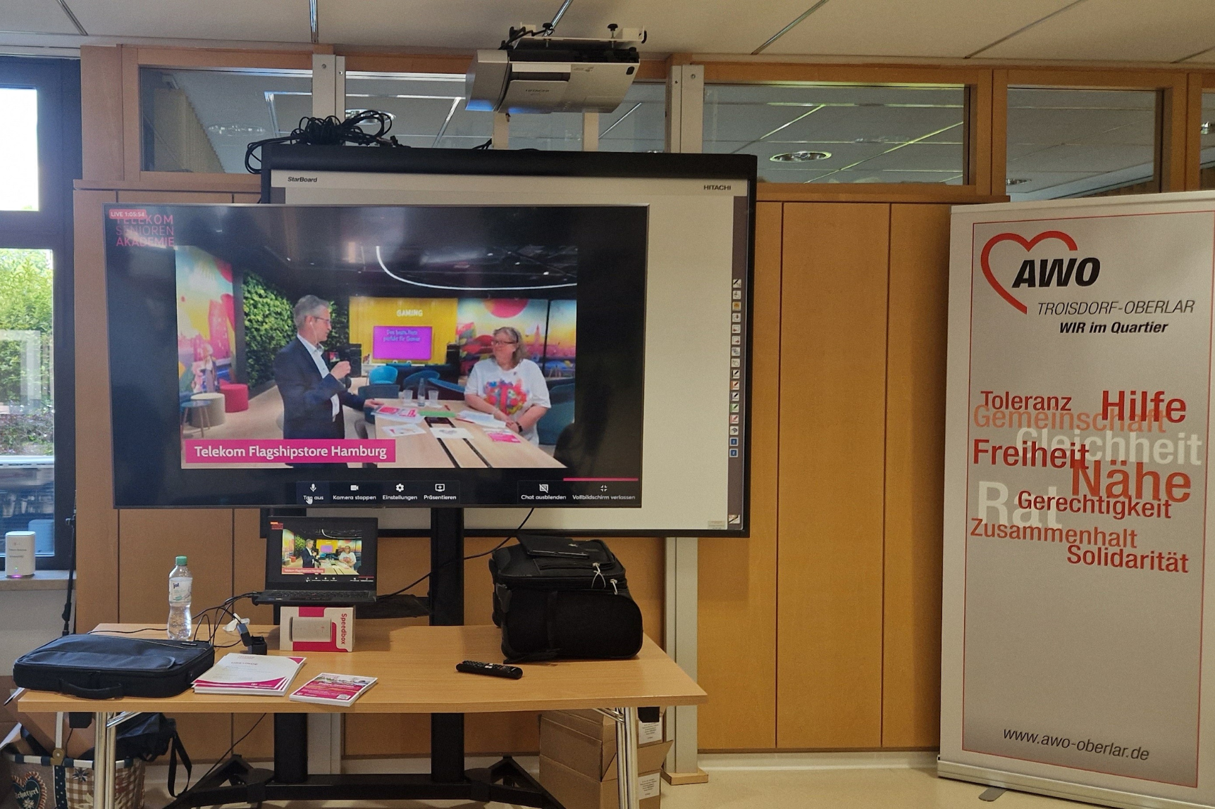 Übertragung der Liveschalte in den Telekom-Store in Hamburg auf einem Fernseherbildschirm, rechts ein Roll-up des AWO Troisdort-Oberlaar.