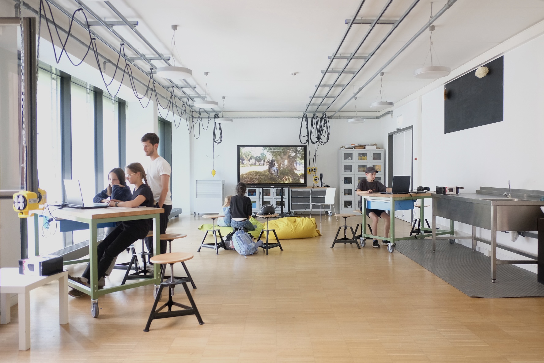 Das Bild zeigt einen modernen Büro- oder Coworking-Raum. Es gibt mehrere Schreibtische mit Stühlen, einige davon sind von Personen besetzt, die an Laptops arbeiten. Der Raum wirkt hell und luftig, mit großen Fenstern auf der linken Seite, durch die natürliches Licht einfällt. Über den Köpfen hängt eine Reihe von Lampen. Auf der rechten Seite gibt es eine Küchenzeile mit Edelstahl-Arbeitsplatten und Geräten. In der Mitte des Raumes befindet sich ein gemütlicher Sitzbereich mit Sitzsäcken und einem kleinen Couchtisch, der einem an der Wand montierten Fernseher gegenübersteht. 