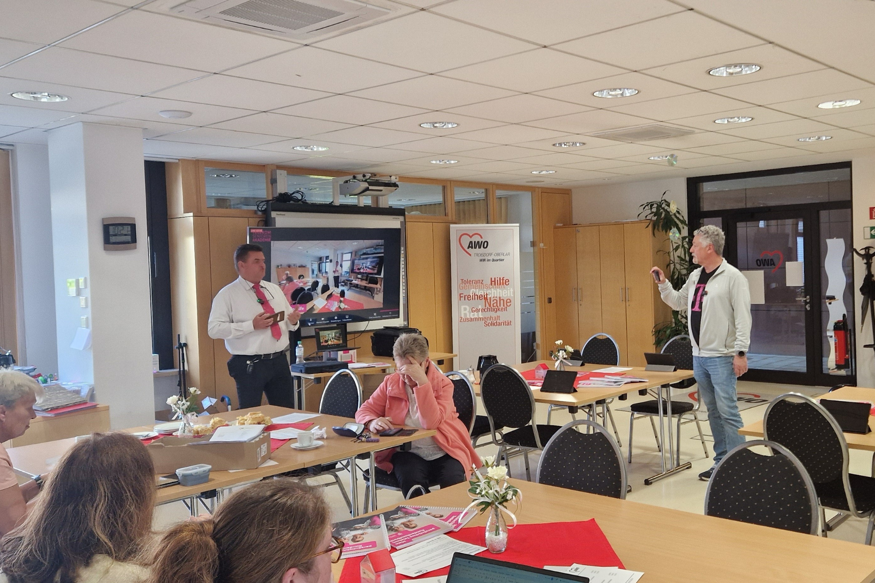 Das Bild zeigt eine Indoor-Veranstaltung mit mehreren Personen. Im Vordergrund befindet sich ein Tisch mit Papieren und einer rosa Broschüre. In der Mitte steht eine Person, die die Gruppe anspricht und ein Mikrofon hält. Rechts steht eine weitere Person neben einem Roll-Up-Banner. Im Hintergrund sind Glastüren zu sehen, die nach draußen führen, sowie ein weiterer Raum gegenüber diesen Türen.