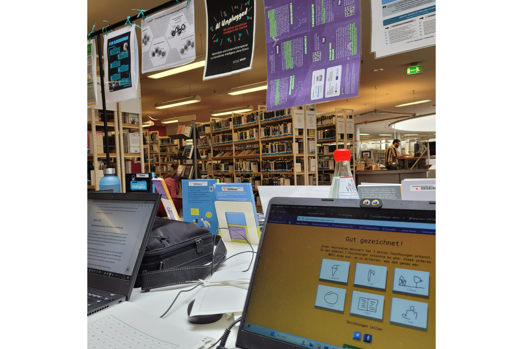 Das Bild zeigt einen Arbeitsplatz in einer Bibliothek mit mehreren geöffneten Laptops auf Schreibtischen. Im Hintergrund sind Bücherregale voller Bücher zu sehen.