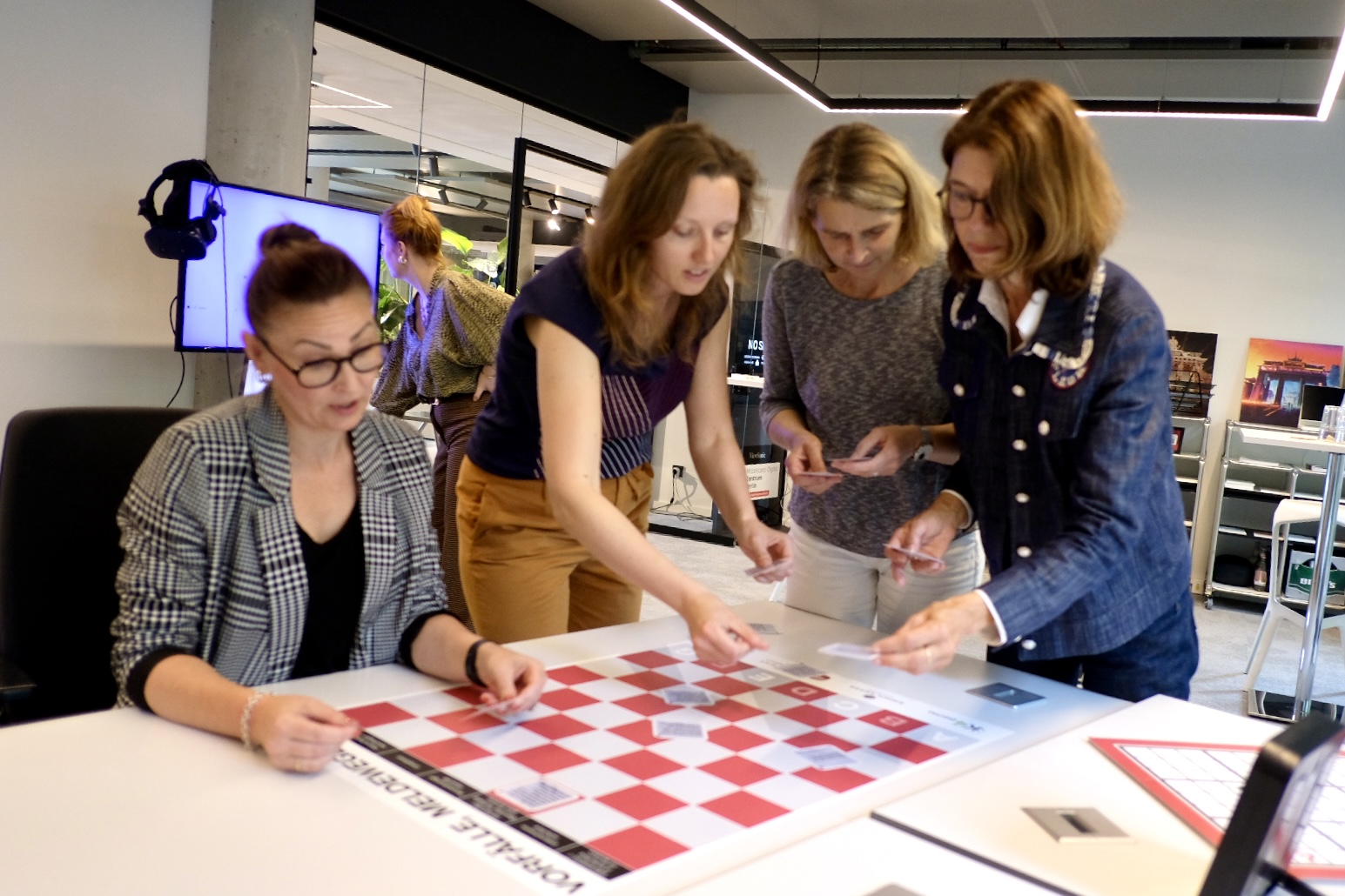 Das Bild zeigt vier Frauen um einen Tisch herum, auf dem ein Spielbrett mit Schachbrettmuster zu sehen ist.