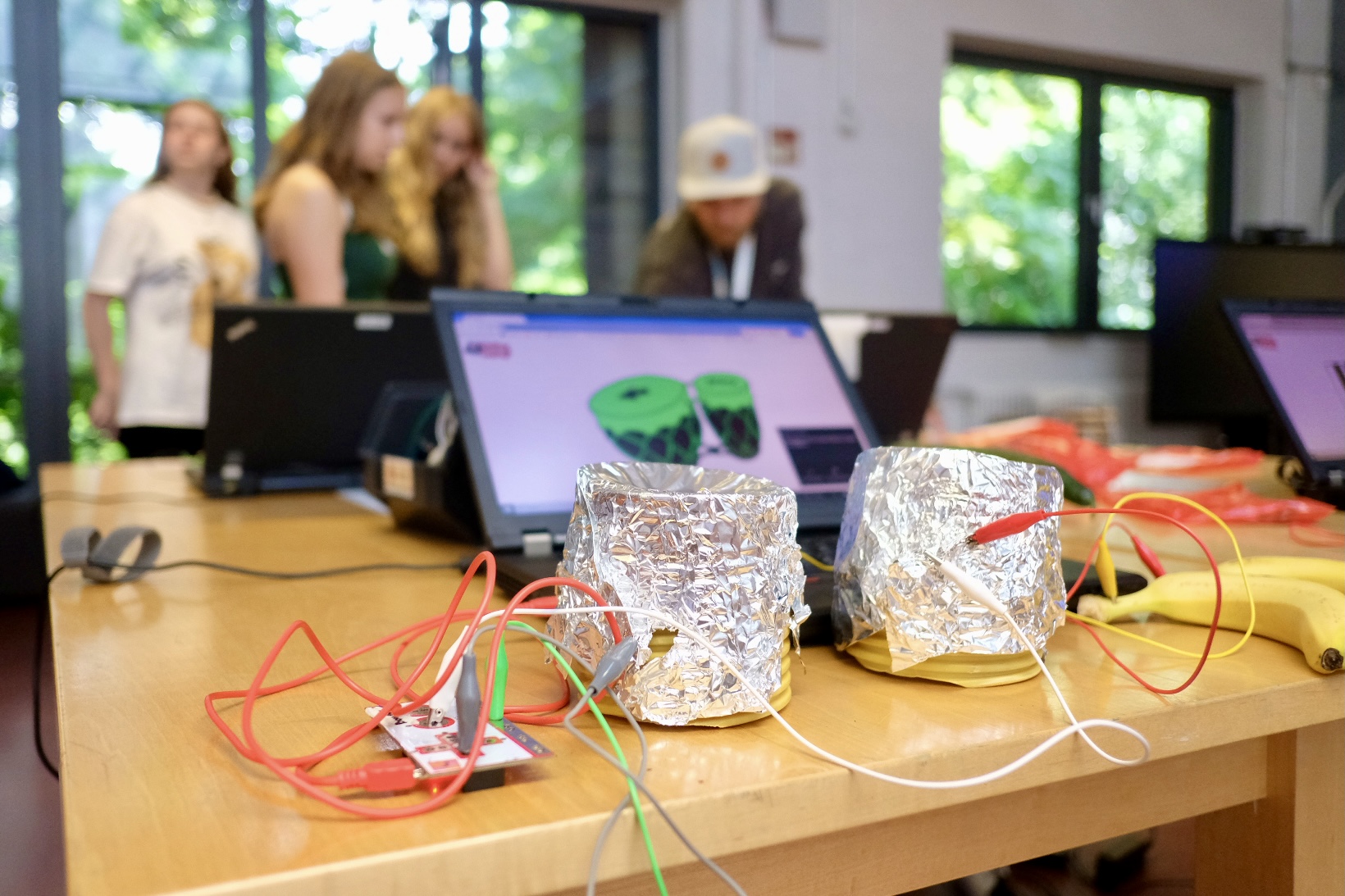 m Vordergrund sind zwei in Aluminiumfolie gewickelte Dosen deutlich auf einem Tisch platziert. Daneben liegen Bananen und bunte Kabel, die um die Dosen verstreut sind. Im Hintergrund sind Laptops zu sehen.