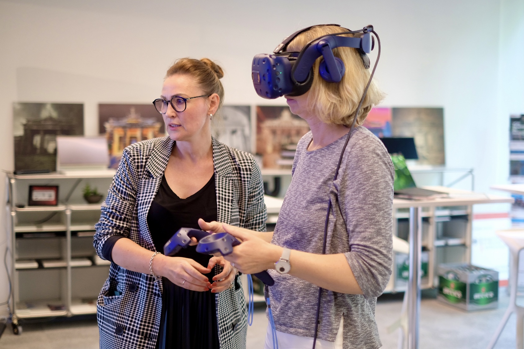 Das Bild zeigt zwei Frauen, wobei eine ein Virtual-Reality-Headset trägt und einen VR-Controller hält. 