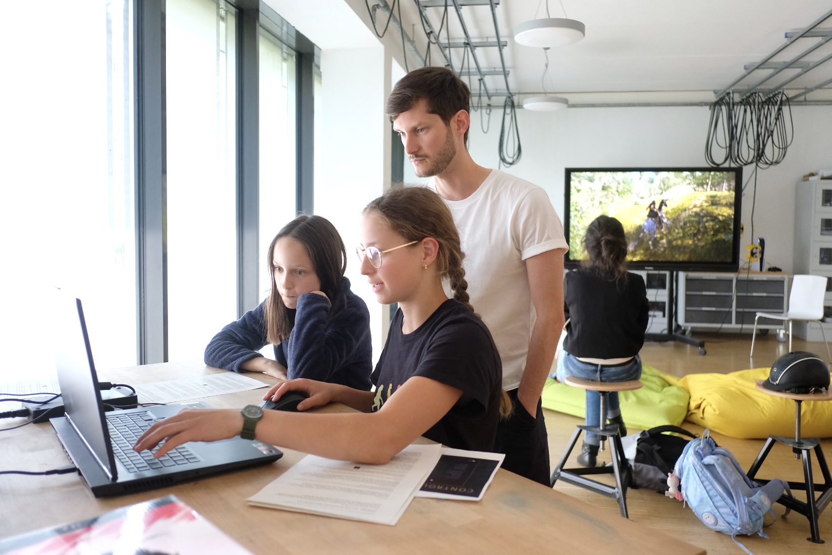 Im Vordergrund sitzen zwei Mädchen vor einem Laptop, hinter ihnen ein junger Mann, der ihnen über die Schulter schaut. Im Hintergrund sitzt eine Person vor einem großen Bildschirm, auf dem ein Computerspiel läuft. 