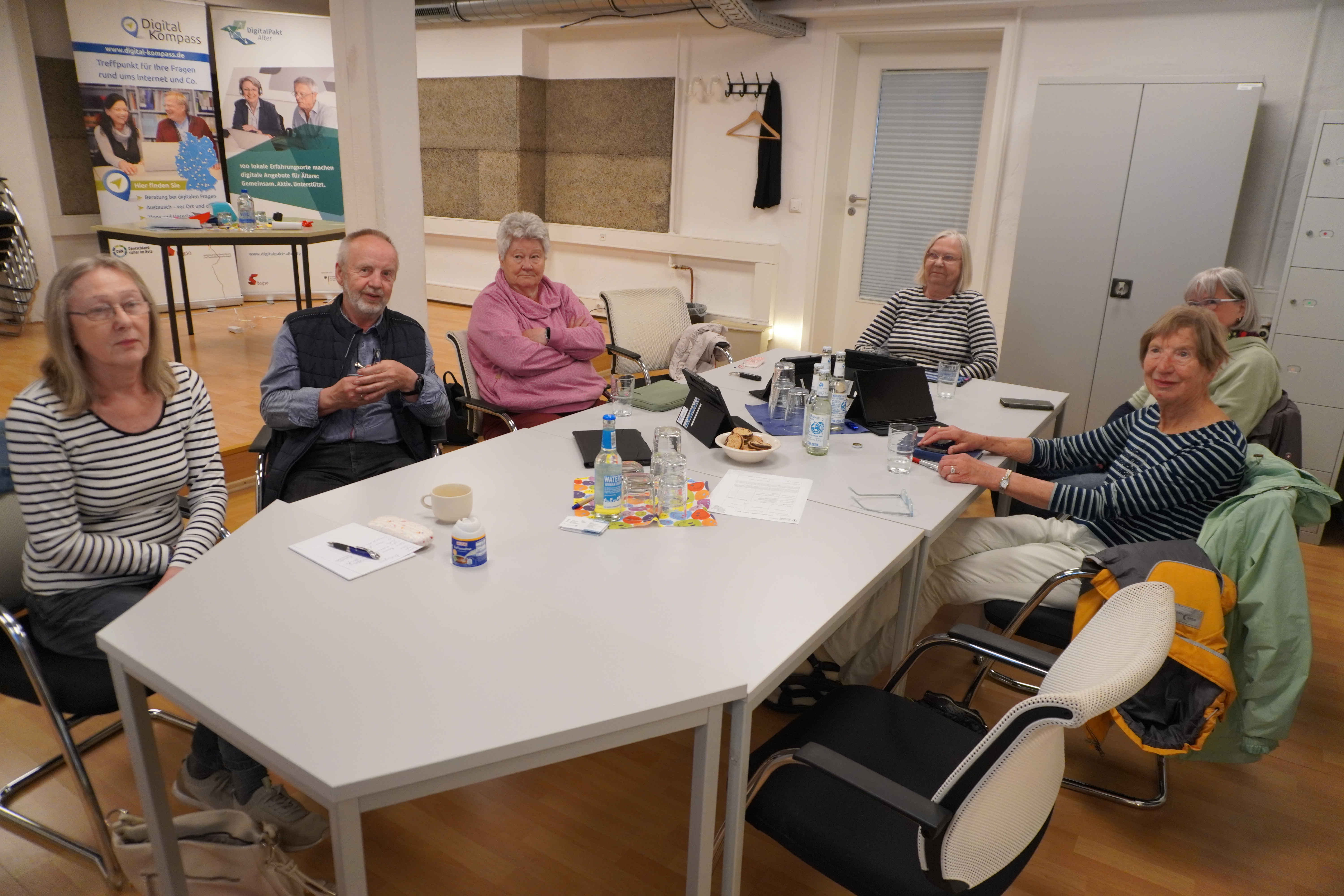 Das Foto zeigt eine Gruppe älterer Menschen, die um einen großen Tisch sitzen. Auf dem Tisch liegen verschiedene Gegenstände wie Papiere, Stifte, Wasserflaschen und Tablets.