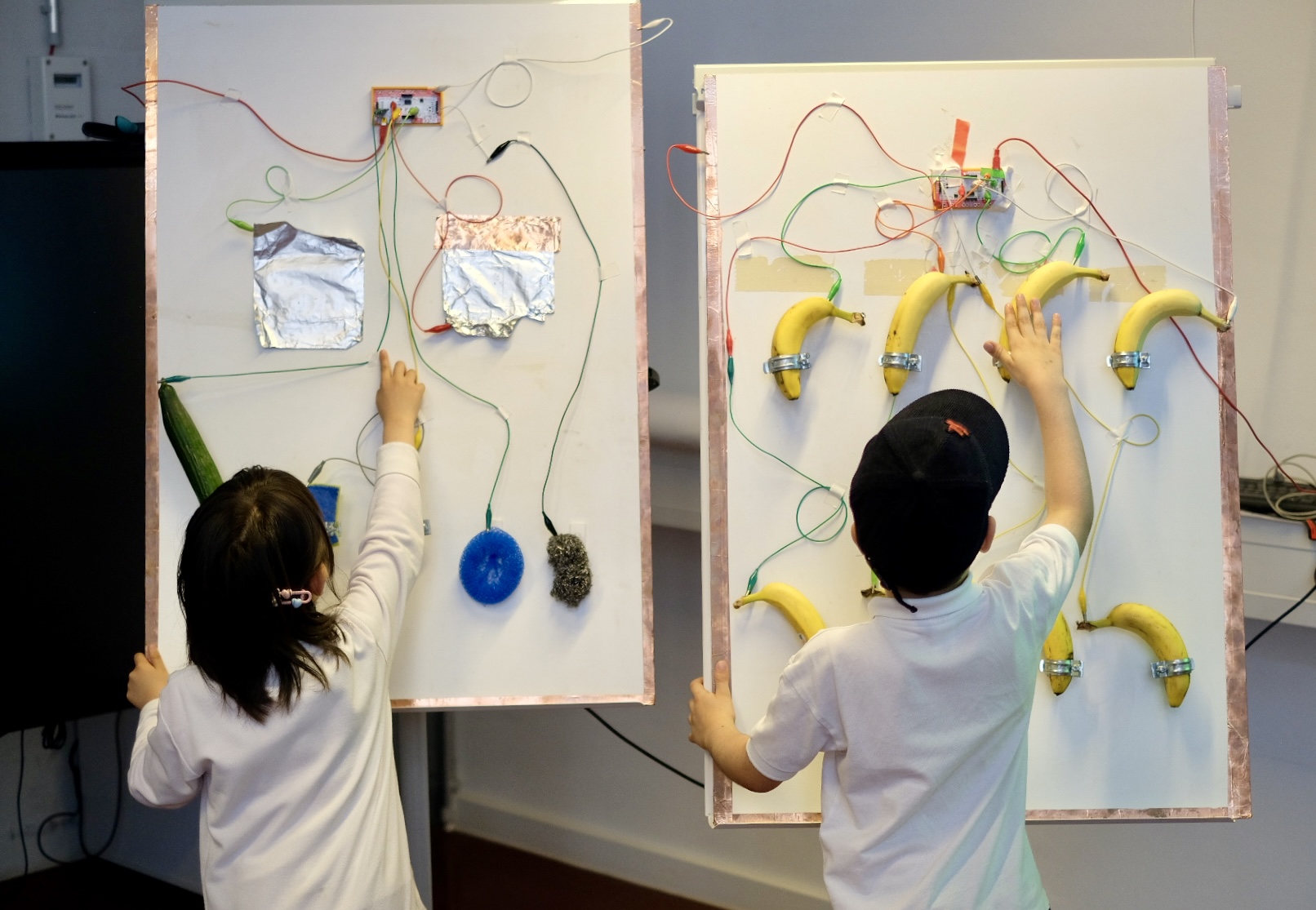 Das Bild zeigt zwei Kinder, die mit einer großen, vertikalen, interaktiven Anzeige interagieren. Die Anzeige umfasst verschiedene Objekte wie Bananen, ein grünes Objekt, das einer Gurke ähnelt, und andere Gegenstände, die mit Drähten mit elektronischen Komponenten auf der Tafel verbunden sind