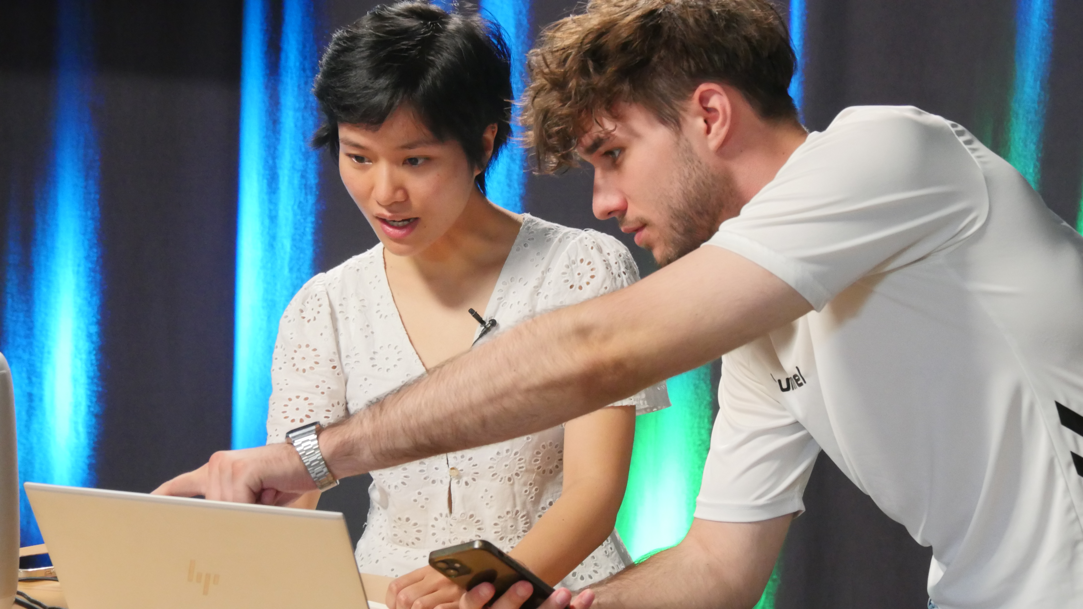 Das Bild zeigt zwei junge Menschen, die gemeinsam an einem Laptop arbeiten. Die eine Person zeigt der anderen auf dem Rechner etwas und streckt dazu den Arm aus. Im Hintergrund sind grün-blaue Lichter zu sehen. 