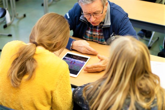 Zwei Mädchen zeigen einem älteren Herren etwas auf dem iPad
