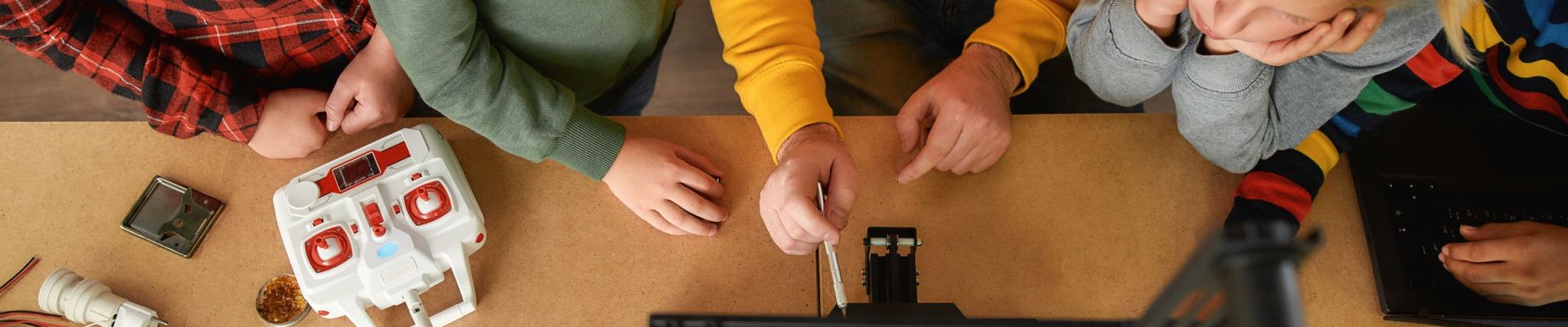 Eine Gruppe von Kindern und ein Erwachsener arbeiten gemeinsam an einem Tisch mit einem 3D-Drucker und elektronischen Bauteilen. Die Kinder schauen interessiert zu, während der Erwachsene etwas am Drucker erklärt oder justiert. Auf dem Tisch liegen verschiedene Werkzeuge, Kabel und ein Fernsteuerungsgerät.