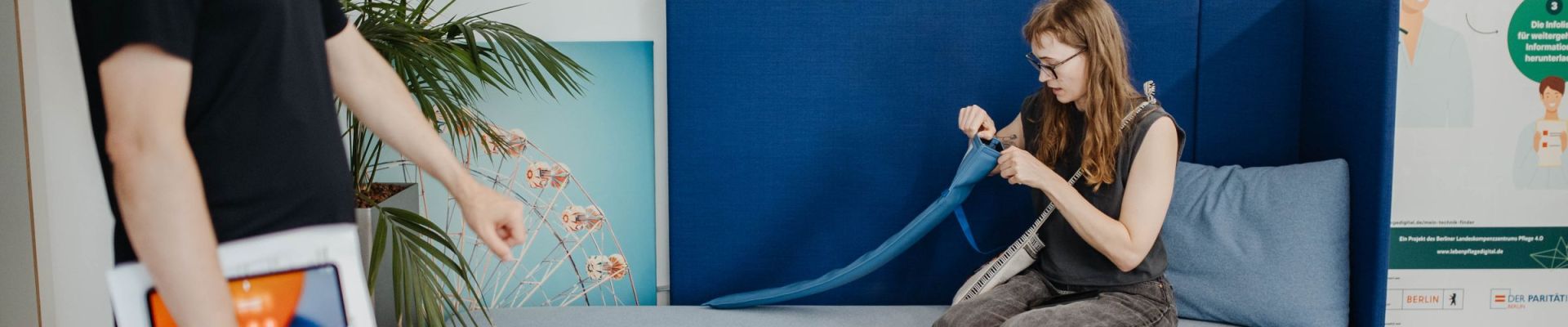 ALT-Text: Eine Frau sitzt auf einem modernen, hellblauen Sofa mit dunkelblauer Rückenlehne und näht konzentriert an einem blauen Stoffband. Neben ihr liegt ein Maßband. Ein Mann im schwarzen T-Shirt hält ein Tablet und geht an ihr vorbei. Im Hintergrund stehen eine Topfpflanze, ein Bild mit einem Riesenrad und ein Aufsteller mit Informationen zu einem Technikprojekt in der Pflege.