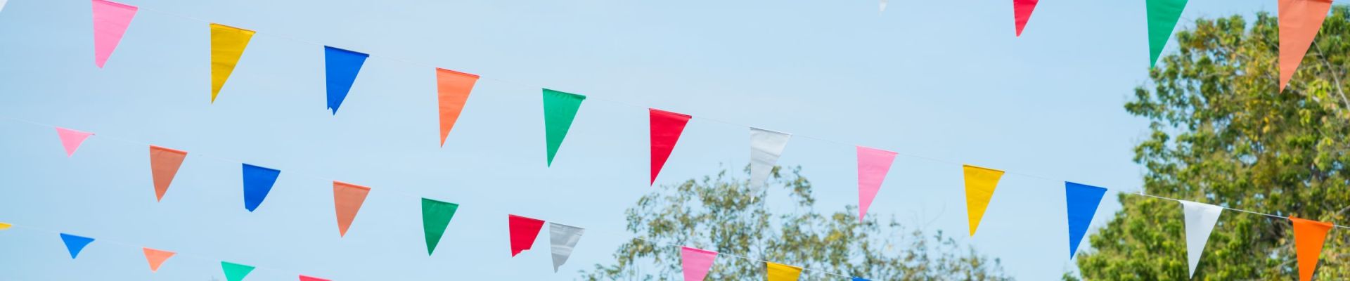 Ein Outdoor-Bild mit vielen bunten Wimpelketten, die zwischen Bäumen und Gebäuden im Hintergrund aufgehängt sind. Die Wimpel sind in verschiedenen Farben wie Blau, Rot, Grün, Gelb und Weiß und hängen vor einem hellblauen Himmel.