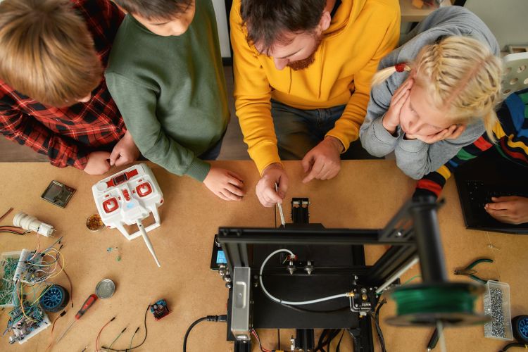 Eine Gruppe von Kindern und ein Erwachsener arbeiten gemeinsam an einem Tisch mit einem 3D-Drucker und elektronischen Bauteilen. Die Kinder schauen interessiert zu, während der Erwachsene etwas am Drucker erklärt oder justiert. Auf dem Tisch liegen verschiedene Werkzeuge, Kabel und ein Fernsteuerungsgerät.