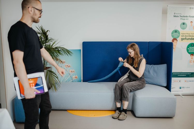 ALT-Text: Eine Frau sitzt auf einem modernen, hellblauen Sofa mit dunkelblauer Rückenlehne und näht konzentriert an einem blauen Stoffband. Neben ihr liegt ein Maßband. Ein Mann im schwarzen T-Shirt hält ein Tablet und geht an ihr vorbei. Im Hintergrund stehen eine Topfpflanze, ein Bild mit einem Riesenrad und ein Aufsteller mit Informationen zu einem Technikprojekt in der Pflege.