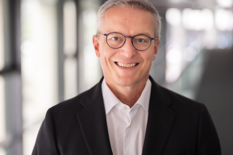 Porträt eines lächelnden Mannes mit kurzen grauen Haaren und runder Brille. Er trägt ein weißes Hemd und ein dunkles Sakko und steht in einem hellen, modern wirkenden Innenraum mit unscharfem Hintergrund.