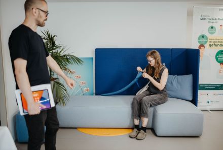 ALT-Text: Eine Frau sitzt auf einem modernen, hellblauen Sofa mit dunkelblauer Rückenlehne und näht konzentriert an einem blauen Stoffband. Neben ihr liegt ein Maßband. Ein Mann im schwarzen T-Shirt hält ein Tablet und geht an ihr vorbei. Im Hintergrund stehen eine Topfpflanze, ein Bild mit einem Riesenrad und ein Aufsteller mit Informationen zu einem Technikprojekt in der Pflege.