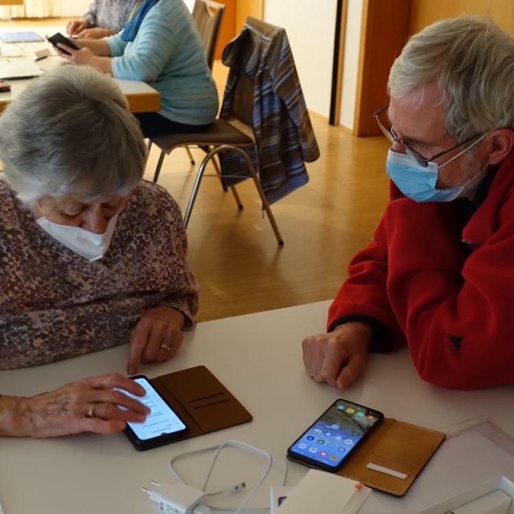 Eine Seniorin beugt sich über ihr Handy; daneben sitzt ihr Pate, ihr beim Bedienen zuschaut.