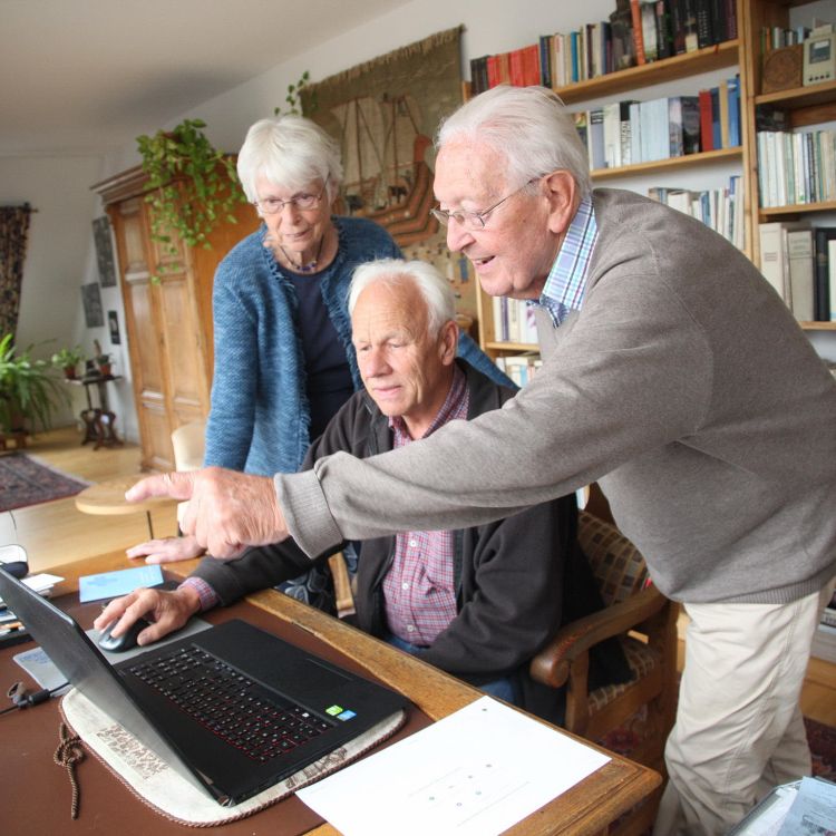 Eine ältere Frau und ein älterer Herr helfen einem Senioren bei der Bedienung eines Laptops.