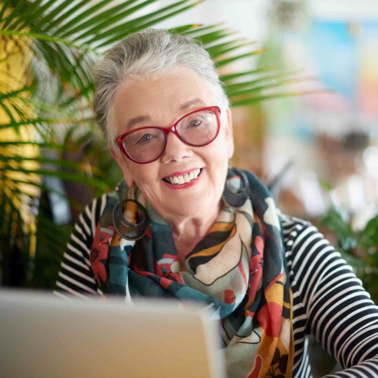 Foto einer älteren Frau mit Brille, die vor einem Laptop sitzt.