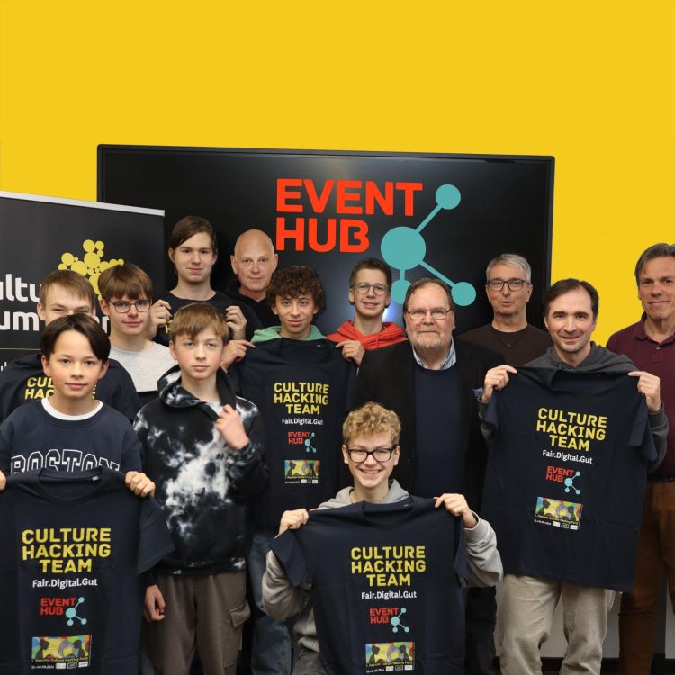 Eine Gruppe von Jugendlichen und Erwachsenen posiert in einem Raum vor einer Leinwand mit der Aufschrift "Event Hub". Einige der Teilnehmenden halten schwarze T-Shirts hoch, auf denen "Future Problem Solving Team" steht. Die Szene vermittelt Teamgeist und Engagement in einem kreativen Projekt.