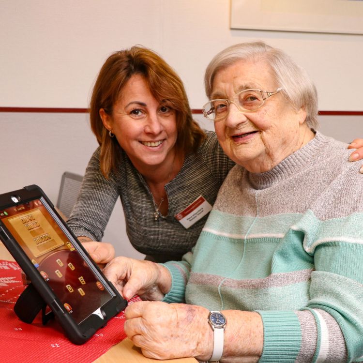 Ein Frau und einer ältere Frau sitzen an einem Tisch, auf dem ein Tablet aufgestellt ist. Auf dem Tablet ist ein Spiel zu sehen.