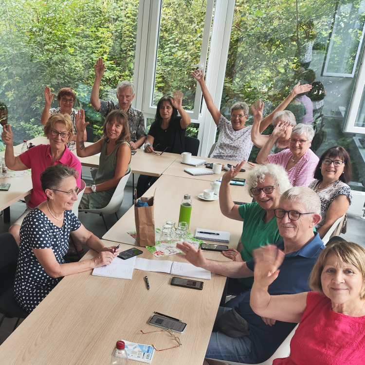 Eine Gruppe von Senioren und Seniorinnen sitzt in einem hellen Raum an mehreren Tischen und lächelt in die Kamera, einige heben die Hände zum Gruß. Die Stimmung wirkt fröhlich und aufgeschlossen, im Vordergrund liegen Schreibmaterialien und Notizen auf den Tischen. Im Hintergrund sind große Fenster zu sehen, durch die viel Tageslicht fällt und Bäume sichtbar sind.