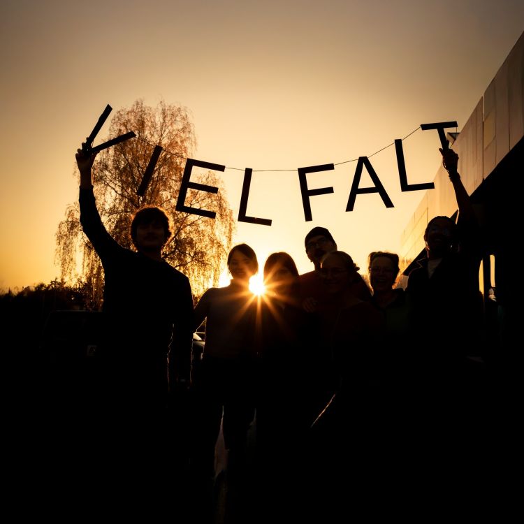 Foto einer Gruppe Menschen, die draußen steht und im Sonnenuntergang eine "Vielfalt"-Girlande hochhält.